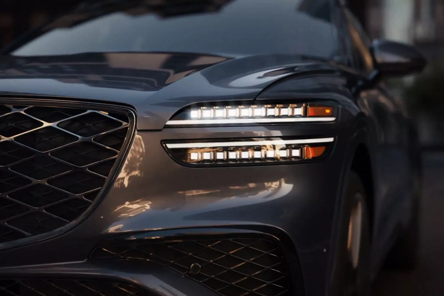 Close-up of a dark grey car's front, showing the grille and illuminated, split LED headlights.